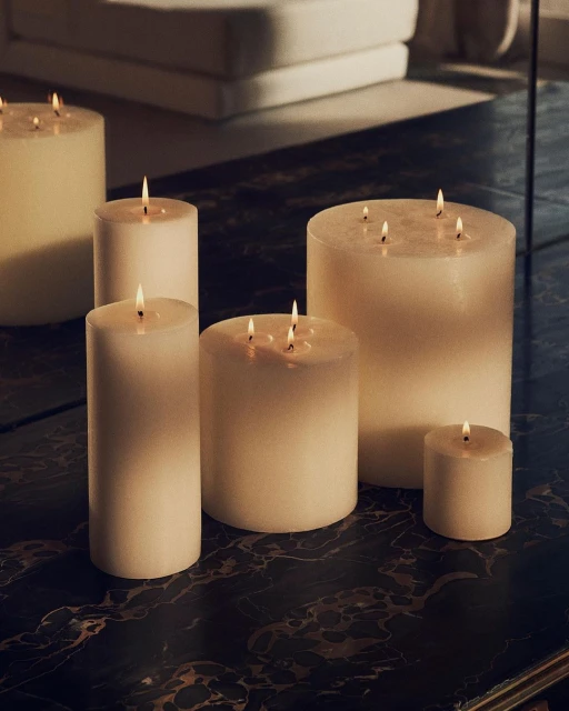 Various sizes of lit candles on a dark marble table, creating a warm and inviting ambiance with a blurred-out background of modern furniture.