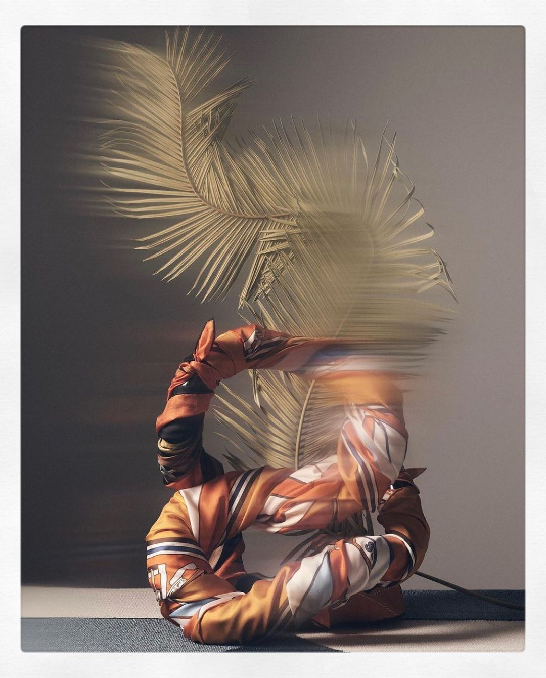 Artistic photo of an orange jacket shaped into a circle with palm fronds, creating an abstract fashion and nature fusion on a gradient background.