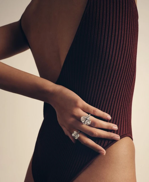 Close-up fashion shot of a slender woman with dark hair, wearing a reddish-brown ribbed bodysuit and diamond rings, against a light background.