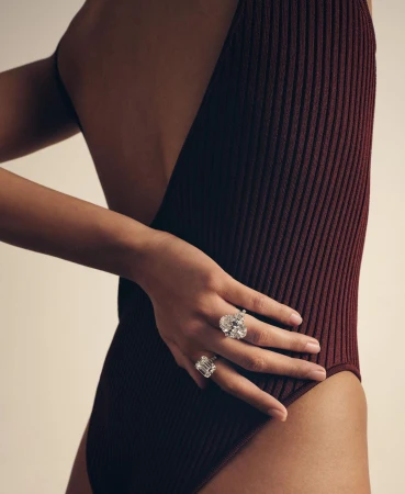 Close-up fashion shot of a slender woman with dark hair, wearing a reddish-brown ribbed bodysuit and diamond rings, against a light background.