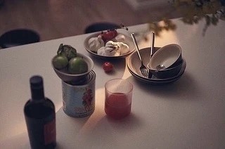 Overhead view of a table with food items including fruit, tomatoes, garlic, and a bottle; warm lighting, soft focus and shallow depth of field.