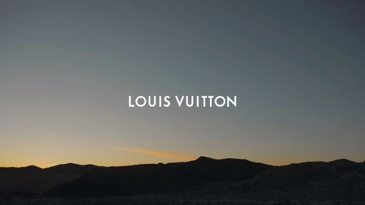 Mountains silhouette and the Louis Vuitton brand text logo in a commercial outdoor shot, during golden hour lighting, for a fashion campaign.