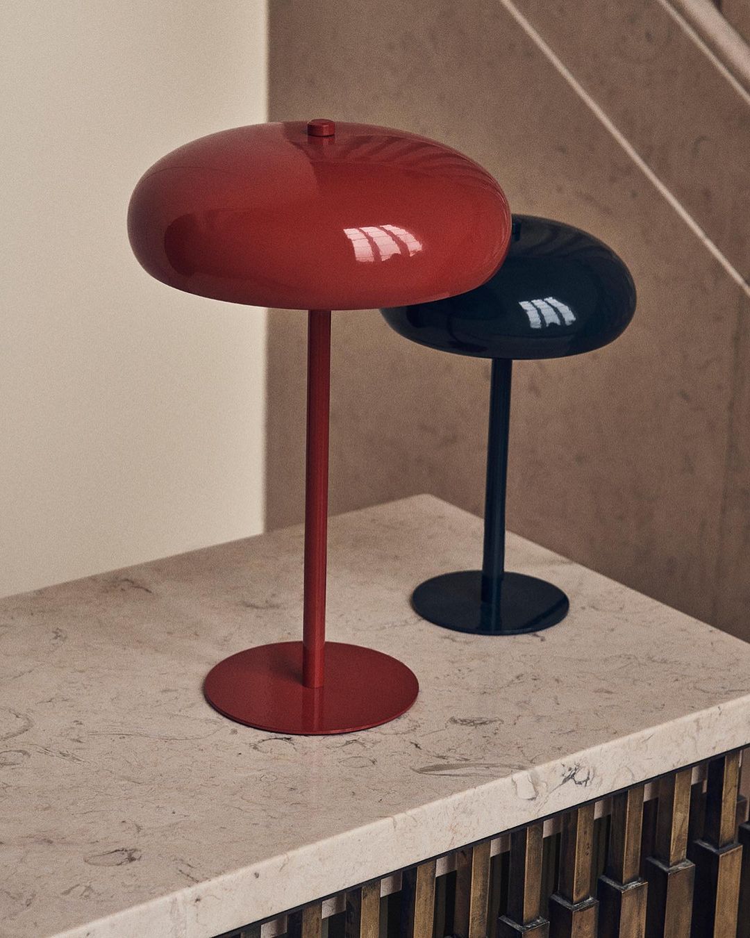 A red lamp and a black lamp sit side-by-side on a light countertop in front of a neutral wall and a staircase with wood trim.