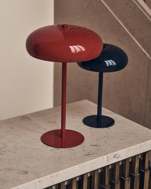 A red lamp and a black lamp sit side-by-side on a light countertop in front of a neutral wall and a staircase with wood trim.