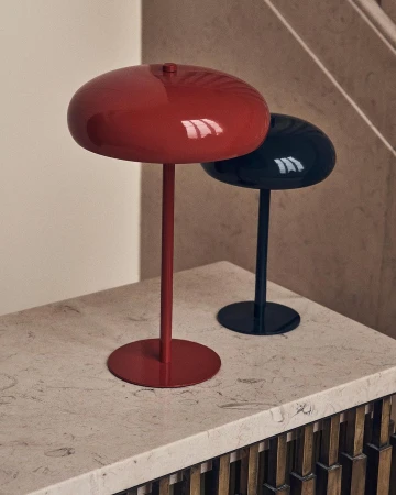 A red lamp and a black lamp sit side-by-side on a light countertop in front of a neutral wall and a staircase with wood trim.