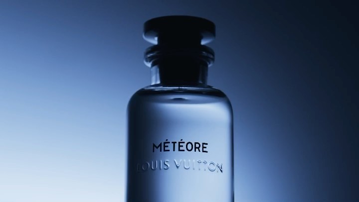 A clear Louis Vuitton cologne bottle, labeled Météore, stands against a gradient blue background, capturing its sleek design and luxury appeal.
