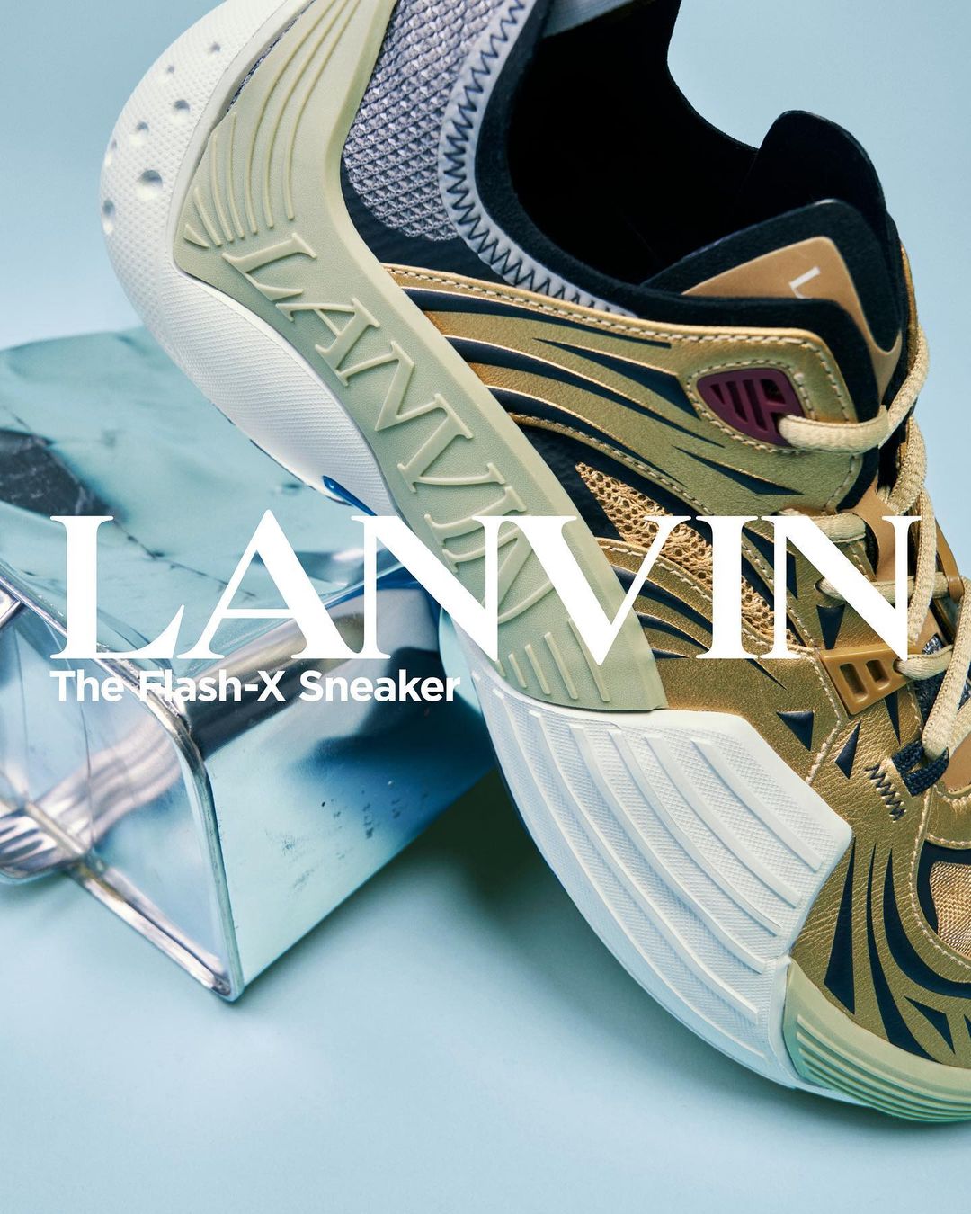 Close-up of a gold and black Lanvin Flash-X sneaker propped on a metallic cube against a light blue background, showcasing its design.