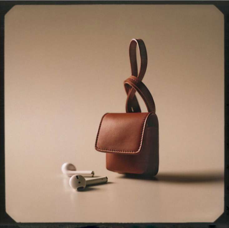 Close-up studio shot of a brown leather wireless earbuds case with a knotted strap, next to white wireless earbuds on a neutral surface.