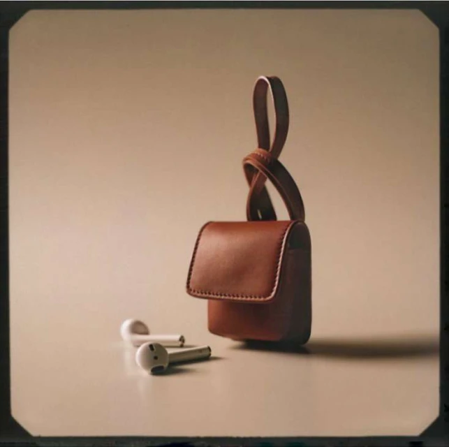 Close-up studio shot of a brown leather wireless earbuds case with a knotted strap, next to white wireless earbuds on a neutral surface.