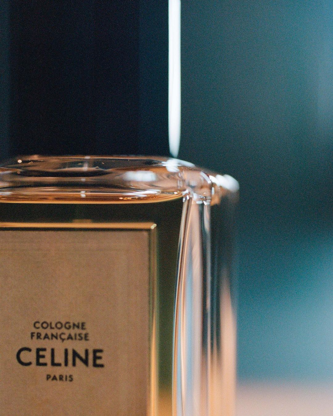 Close-up of a clear glass Celine perfume bottle with a black cap and gold label against a dark blue background.