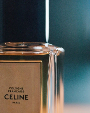 Close-up of a clear glass Celine perfume bottle with a black cap and gold label against a dark blue background.