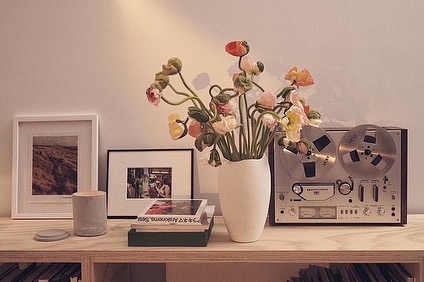 Still life of a vintage reel-to-reel tape recorder next to a vase of flowers, framed art, books, and decorative objects on a wooden shelf.