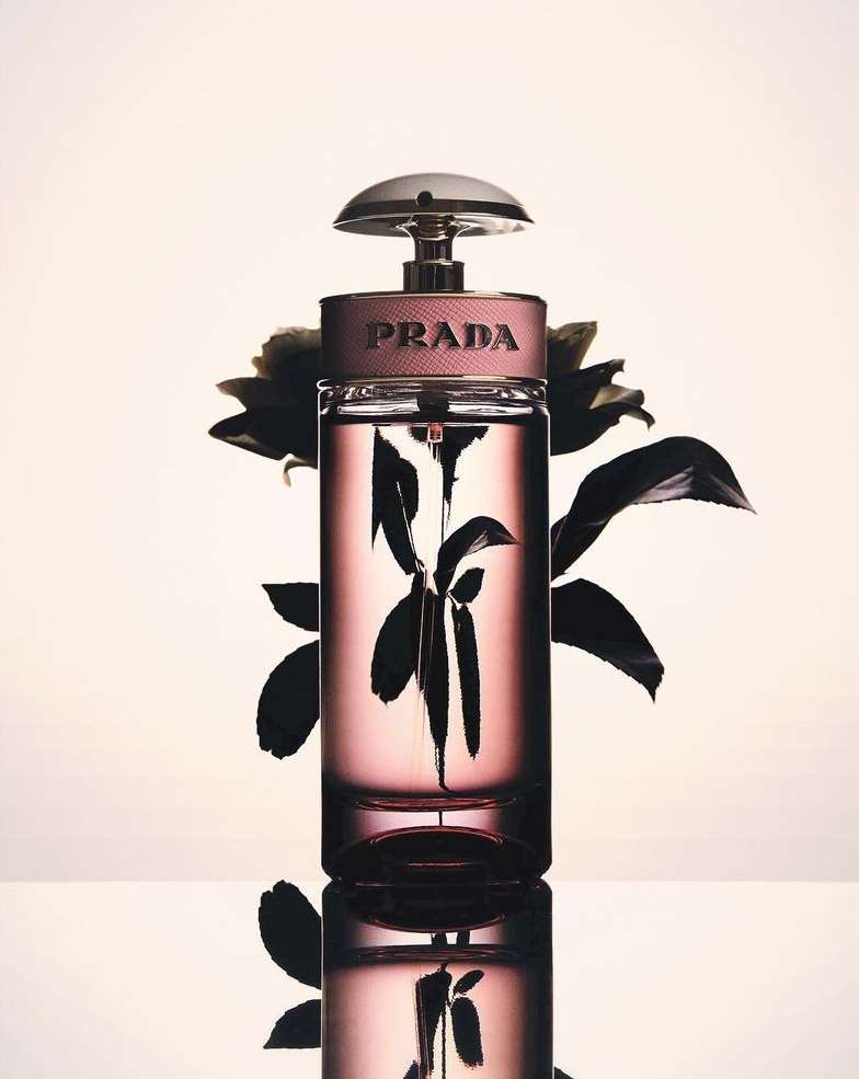 A close-up studio shot features a Prada fragrance bottle with a pink tint, displayed on a reflective surface against a light background.
