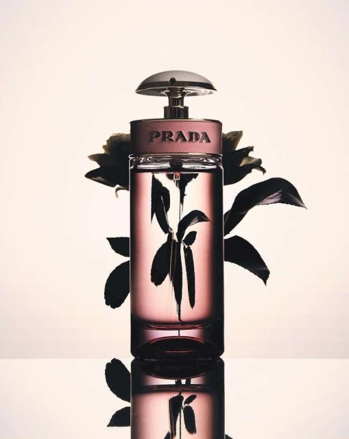 A close-up studio shot features a Prada fragrance bottle with a pink tint, displayed on a reflective surface against a light background.