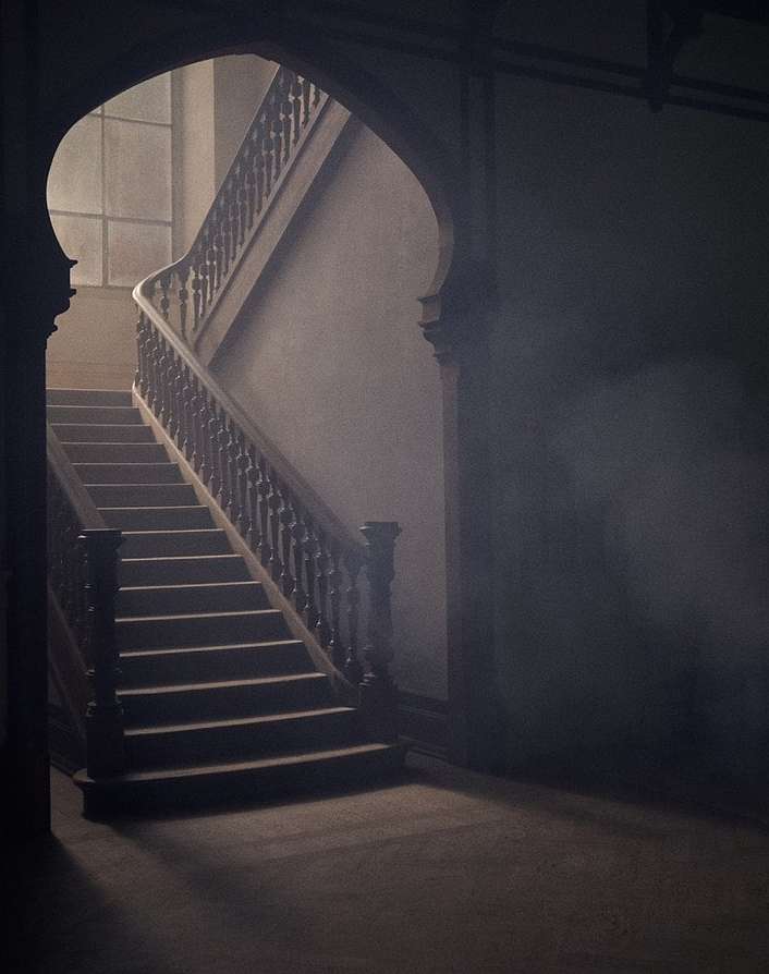 Ornate staircase with carved wooden railings winds upward, bathed in soft, diffused light filtering through a high window in a vintage interior.