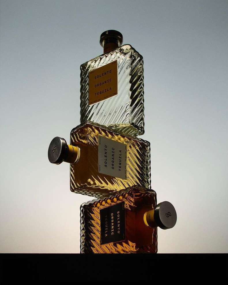 Three square clear glass bottles of organic tequila stacked on a dark surface against a light blue and white gradient background.
