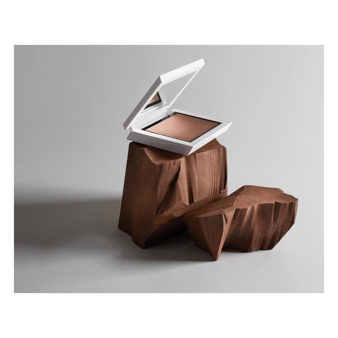 Powder foundation compact in a white case, displayed on carved brown wooden blocks against a gray background, product photography.