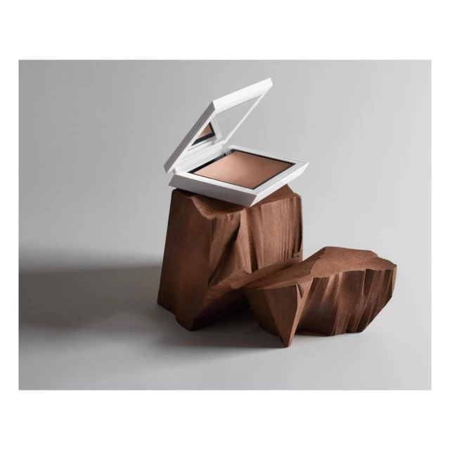 Powder foundation compact in a white case, displayed on carved brown wooden blocks against a gray background, product photography.