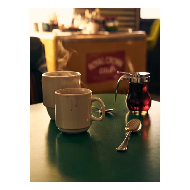 Still life of two mugs, a spoon, and a syrup dispenser on a green table, set against a blurred background of a diner interior.
