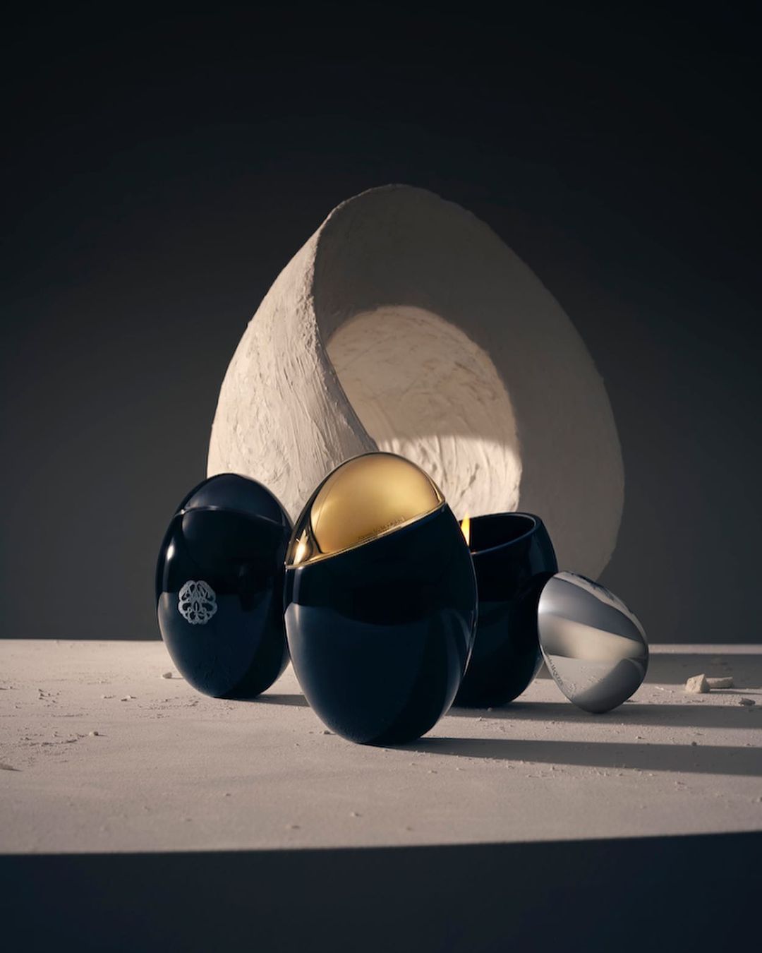 Four black and silver cosmetic containers with an ovoid shape are arranged on a cream surface, with a large, textured semi-circle in the backdrop.
