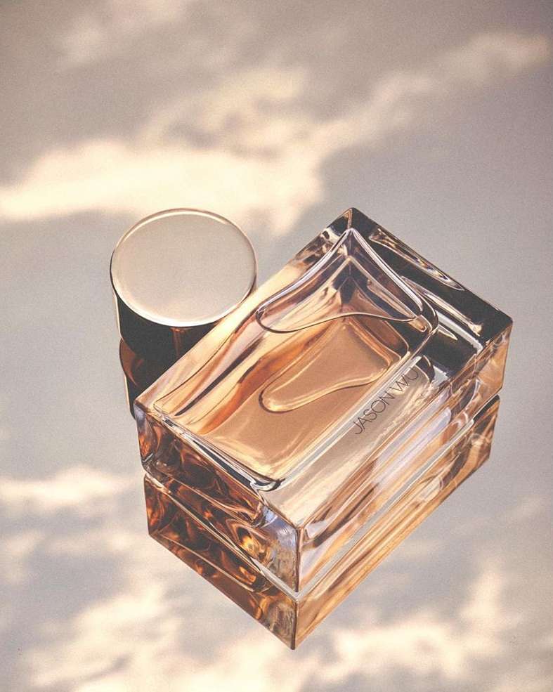 Crystal perfume bottle by Jason Wu with a lid is displayed on a reflective surface in a studio with soft lighting and cloud backdrop.