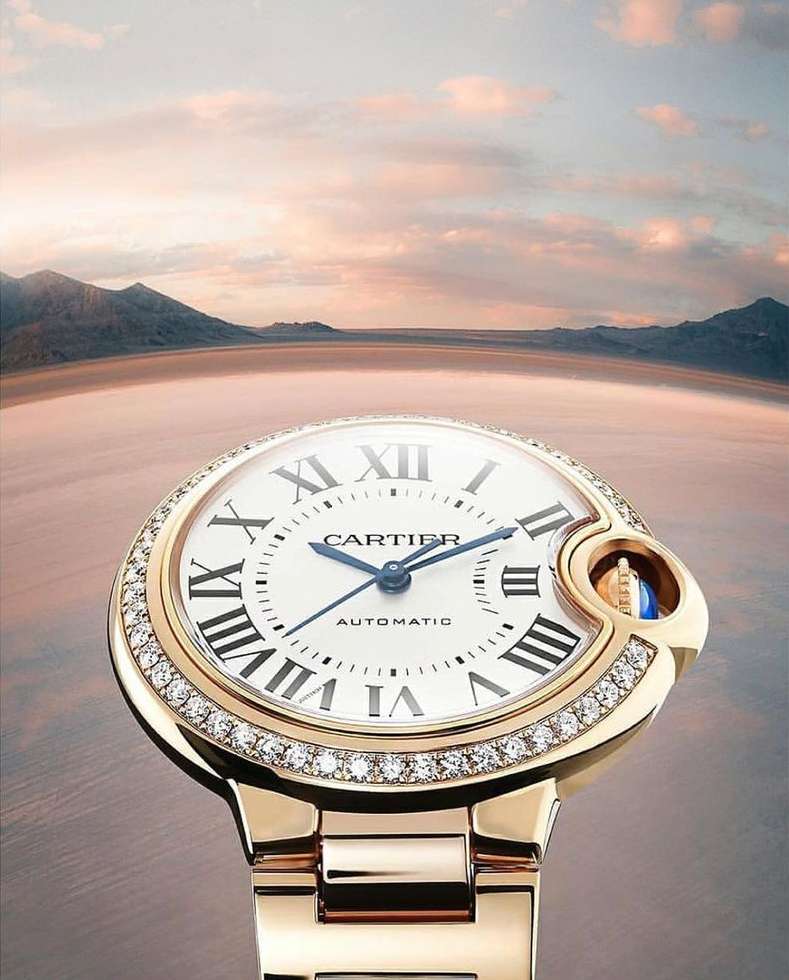 Close-up of a gold Cartier watch with a diamond bezel on a blurred background of a salt flat and distant mountains at sunset.