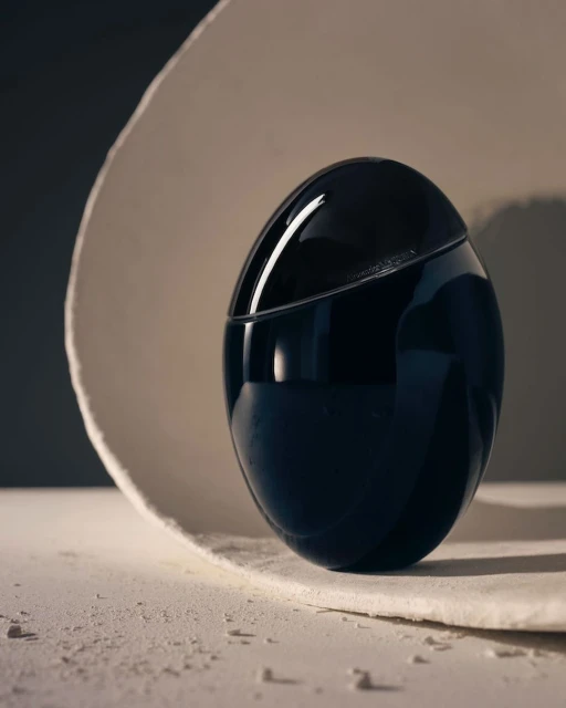 Studio shot of a black, egg-shaped perfume bottle on a textured white surface with a curved white backdrop in soft lighting.