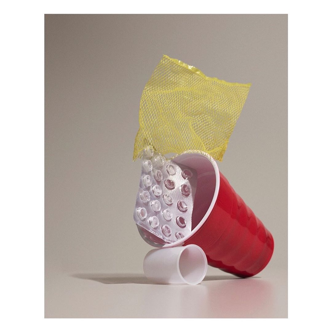 Still life of a red plastic cup, a blister pack with round pills, yellow mesh fabric, and a white plastic ring on a white surface.