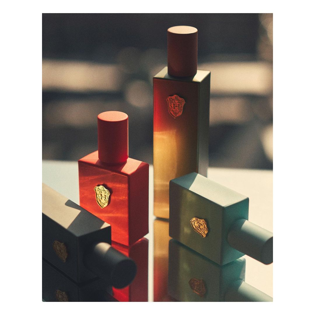 Three geometrically-shaped perfume bottles with unique red, gold, and teal color variations, and golden emblem, displayed on reflective surface.
