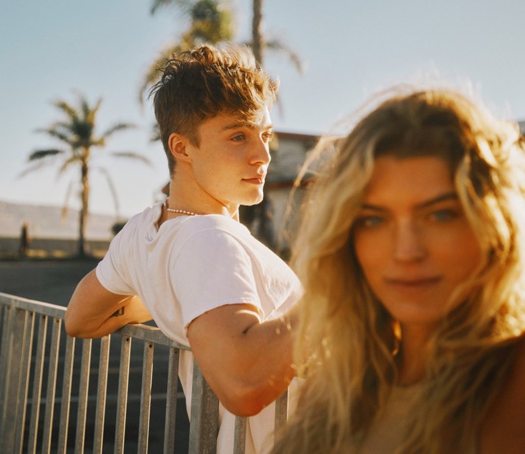 A young couple poses outdoors on a sunny day; a man with short brown hair leans against a railing, with a woman in the foreground.
