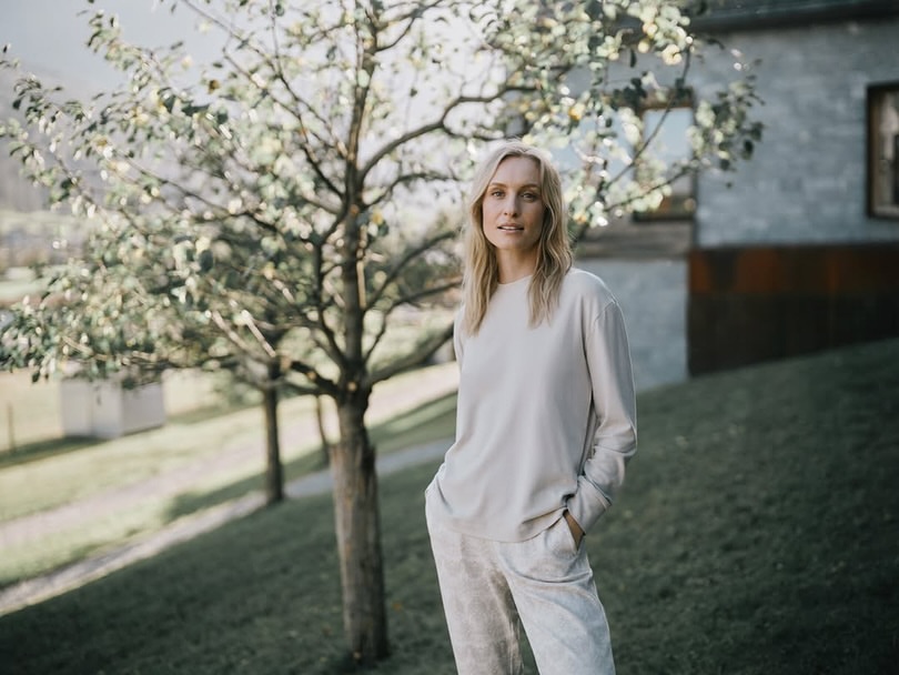 Blond woman poses in a casual outfit near a tree in a bright, outdoor setting with a glimpse of a house in the background.
