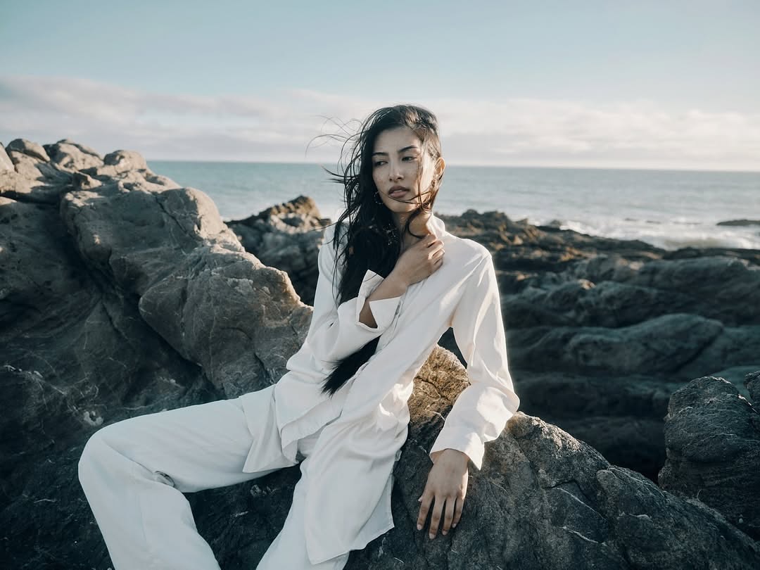 Woman in white clothes poses on rocks by the ocean under a bright sky, exuding a sense of serene fashion and natural beauty.