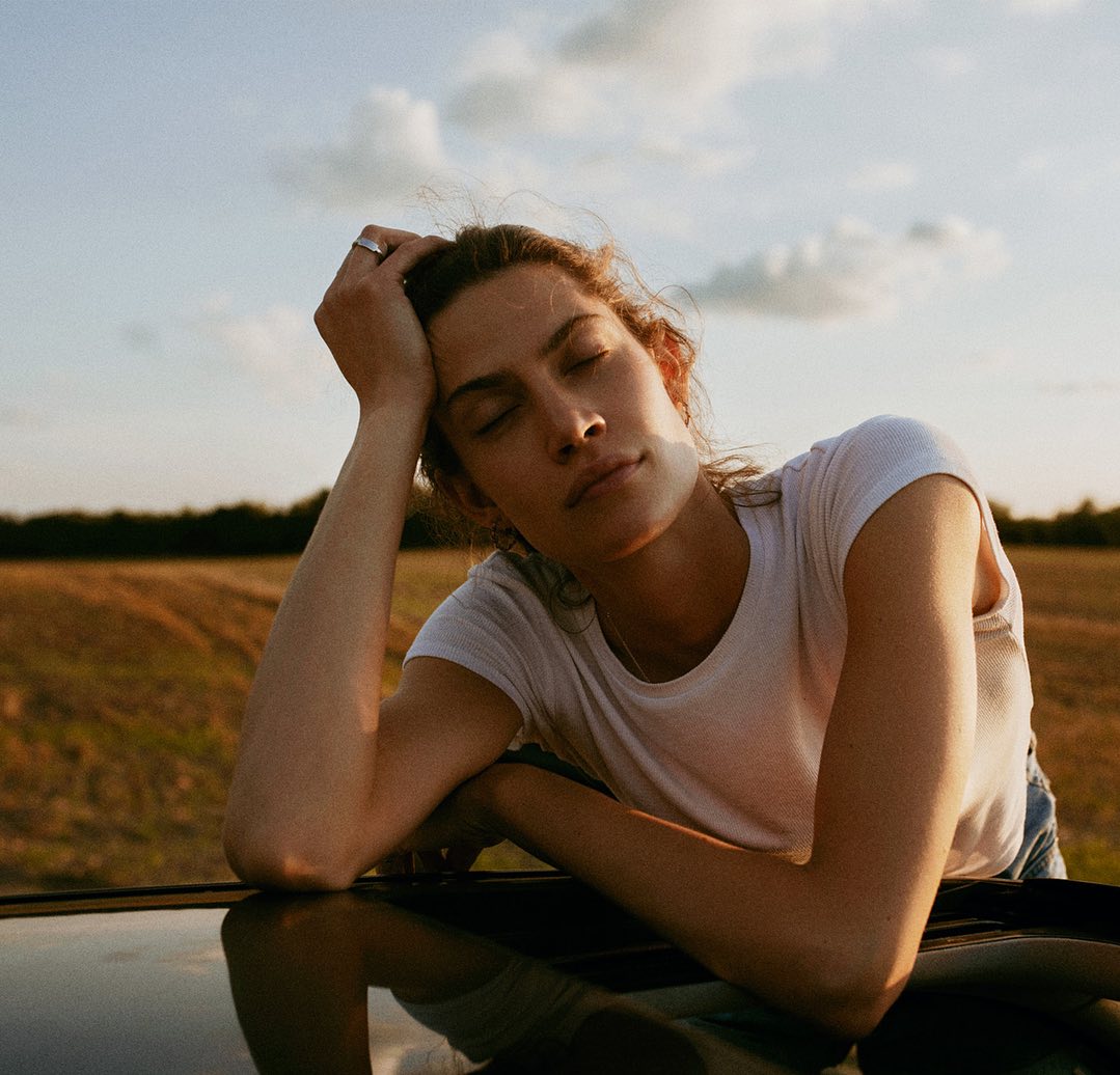Portrait of an adult woman with closed eyes resting on a car roof at sunset, with a farm field and cloudy sky in the background.
