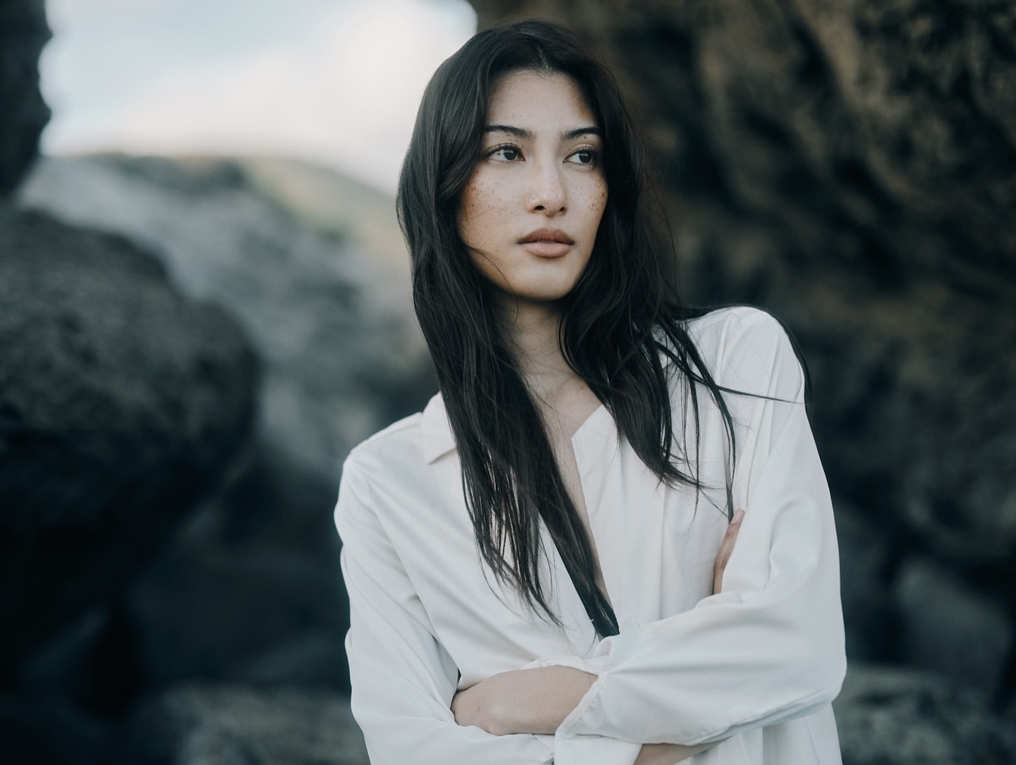 Portrait of a young woman with long dark hair wearing an unbuttoned white shirt, posing outdoors with a rock formation background.