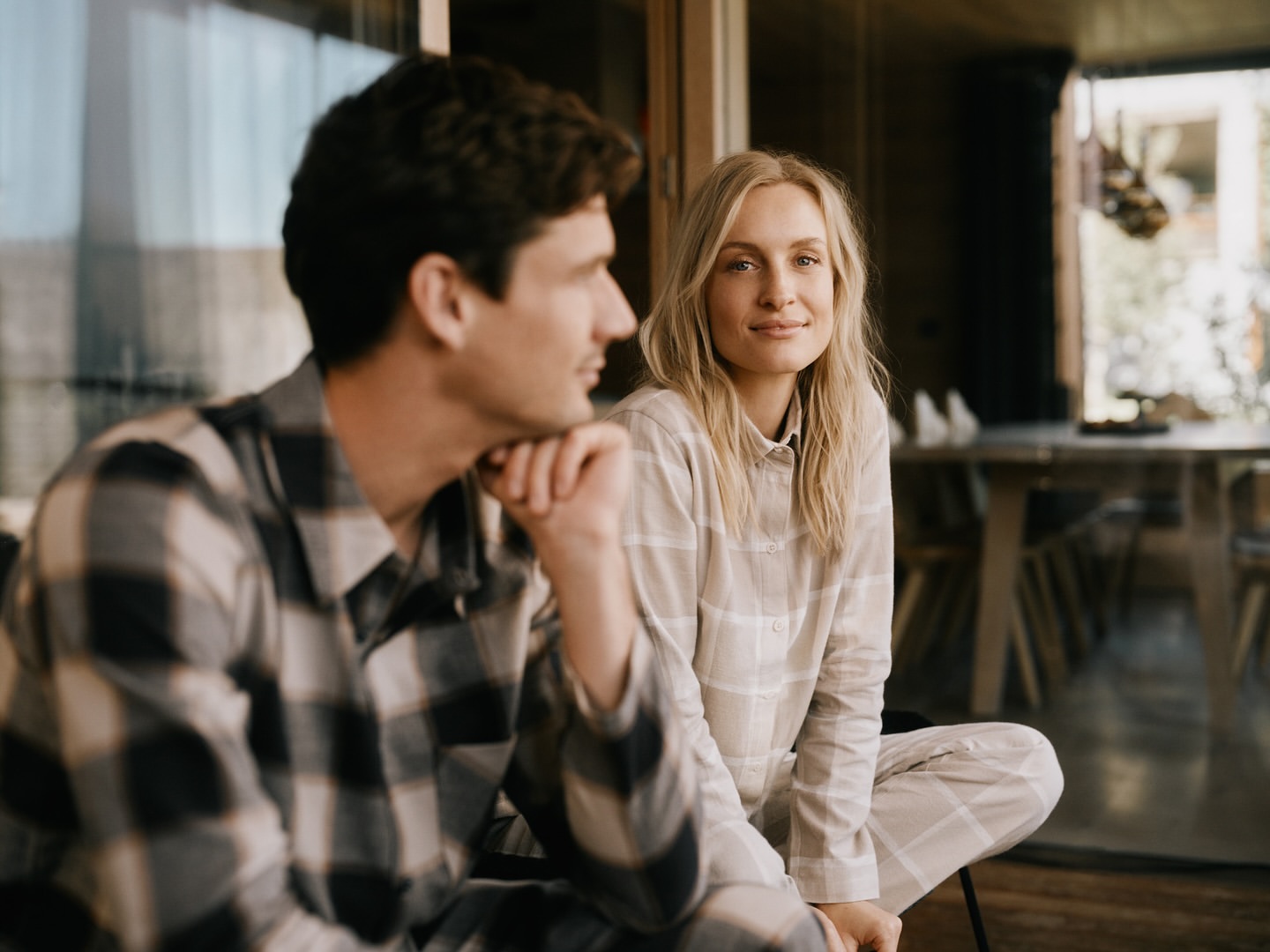 A young man and woman, a couple, are indoors in matching plaid pajamas. They are relaxing in natural light with a window view.
