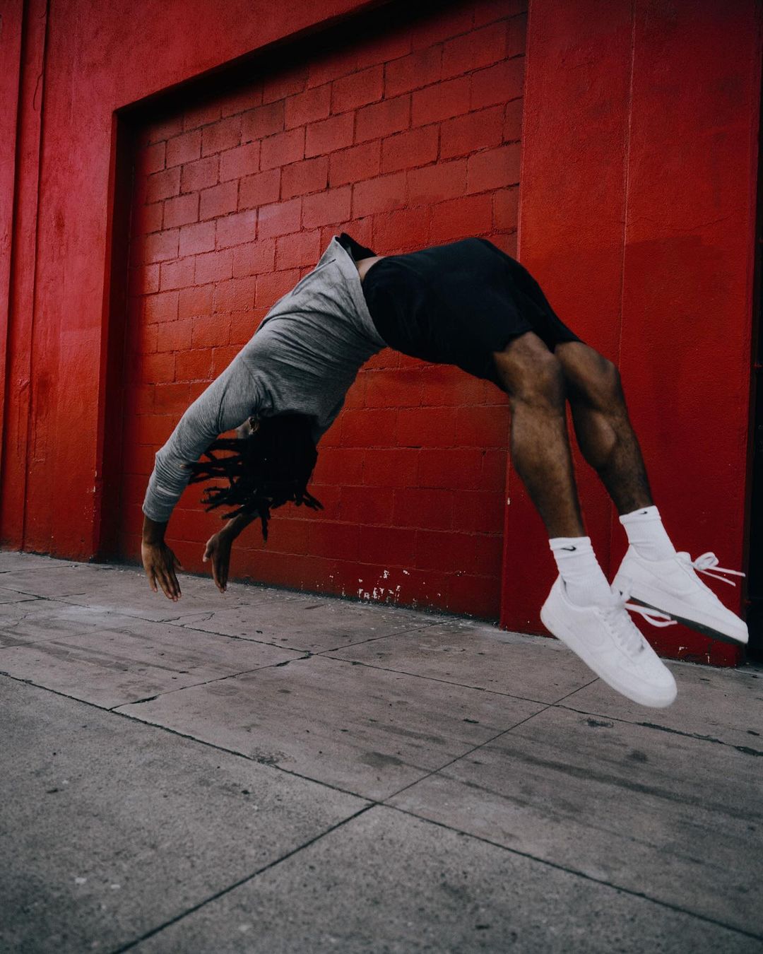 Athletic African-American man in gray shirt and black shorts performs a backflip on city sidewalk with a red brick wall background.