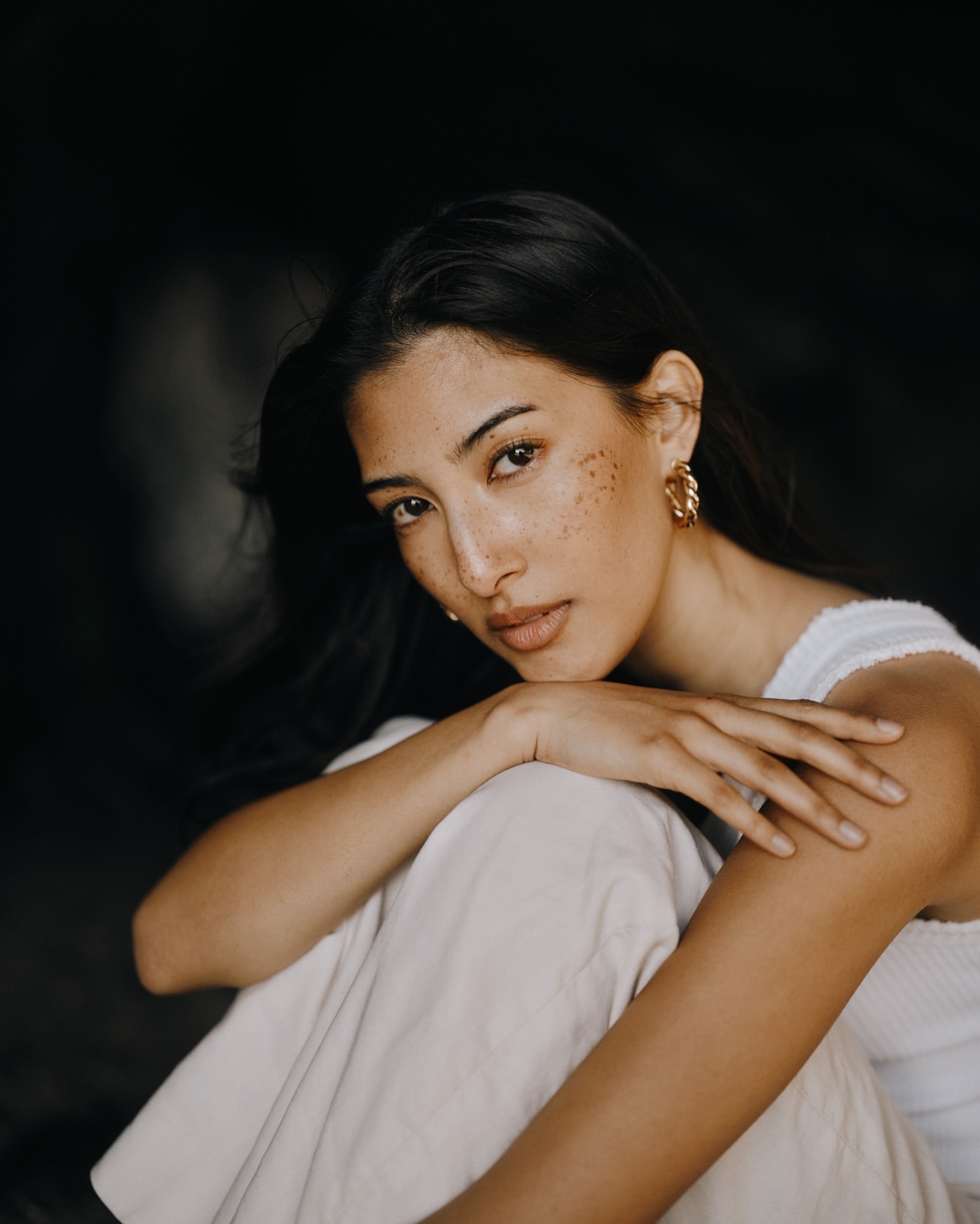 Portrait of a woman with freckles wearing white tank top and beige pants, sitting in front of a dark background with arms resting on knees.