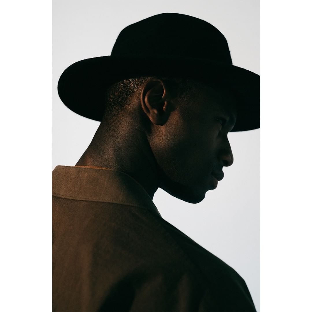 Portrait of a Black man in a fedora and brown jacket against a white backdrop, captured in a medium shot with low-key lighting.