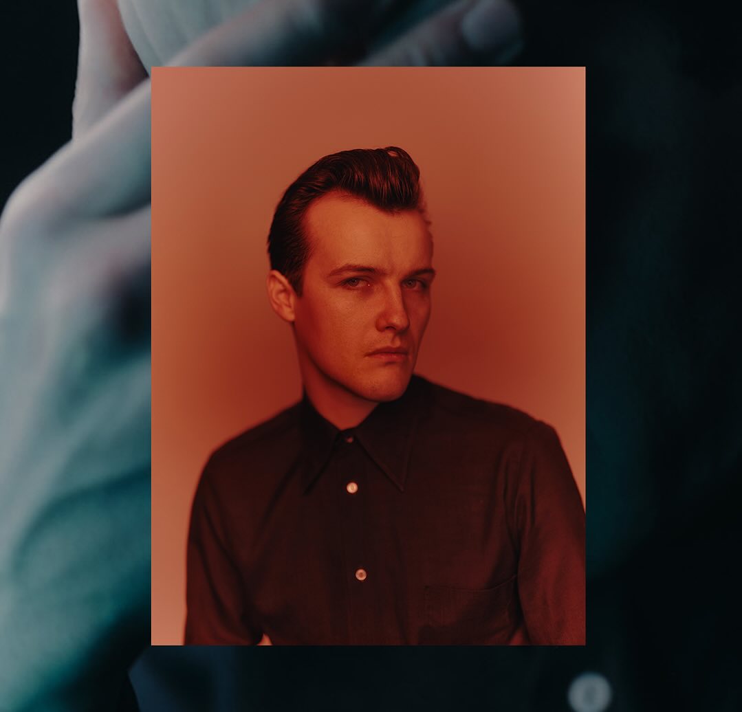 Headshot of a young adult male with dark hair and a collared shirt against a warm-toned studio background with soft, diffused lighting.