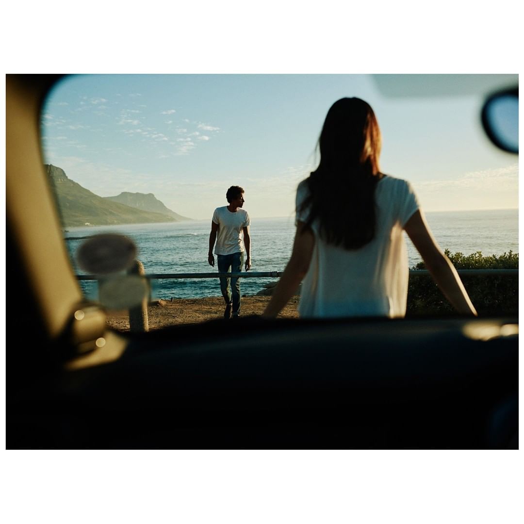 A couple stands near a car overlooking the ocean and mountains. The woman faces forward, and the man walks away from her toward the ocean.