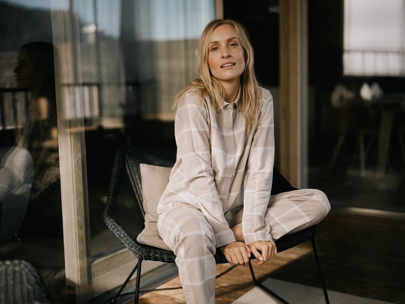 Woman with long blonde hair in neutral plaid pajama set sits in woven chair on sunlit patio with soft focus background.