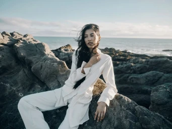 A woman in white clothing seated on dark rocky coastal rocks by the ocean with wind blowing through her dark hair.