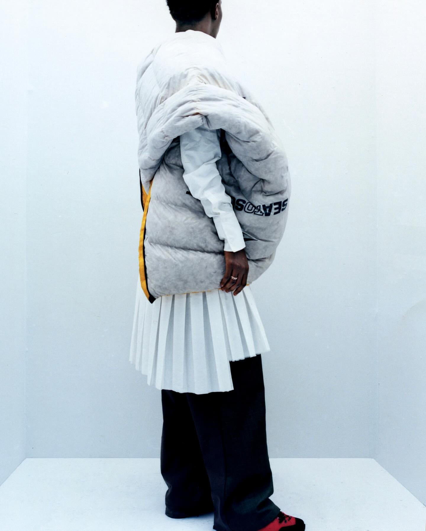 Fashionable person stands in a corner wearing a white shirt, pleated skirt, puffer coat, wide-legged black trousers, and red and black shoes.