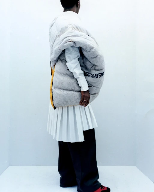 Fashionable person stands in a corner wearing a white shirt, pleated skirt, puffer coat, wide-legged black trousers, and red and black shoes.