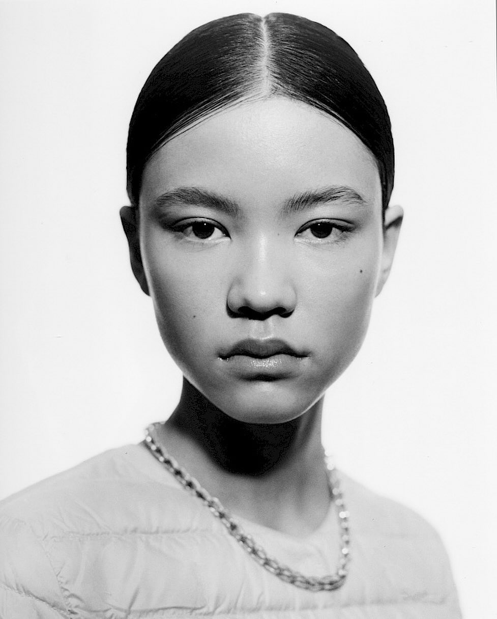 Close-up portrait of an Asian woman with sleek dark hair, wearing a pale yellow quilted jacket and a silver chain necklace, on white.