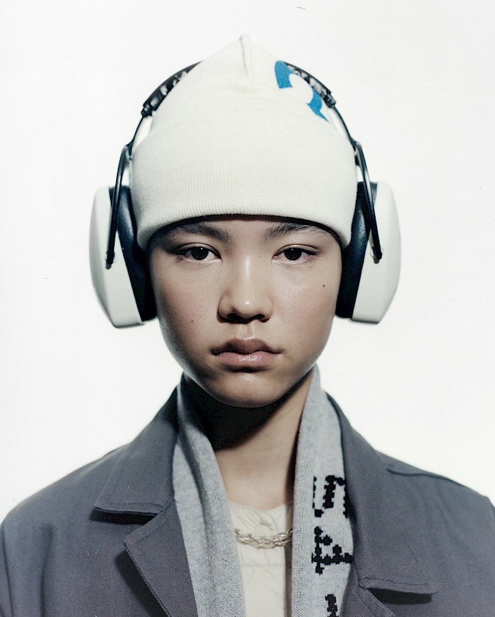 Portrait of an Asian person wearing a white beanie, noise-canceling headphones, gray scarf, and gray blazer against a white backdrop.