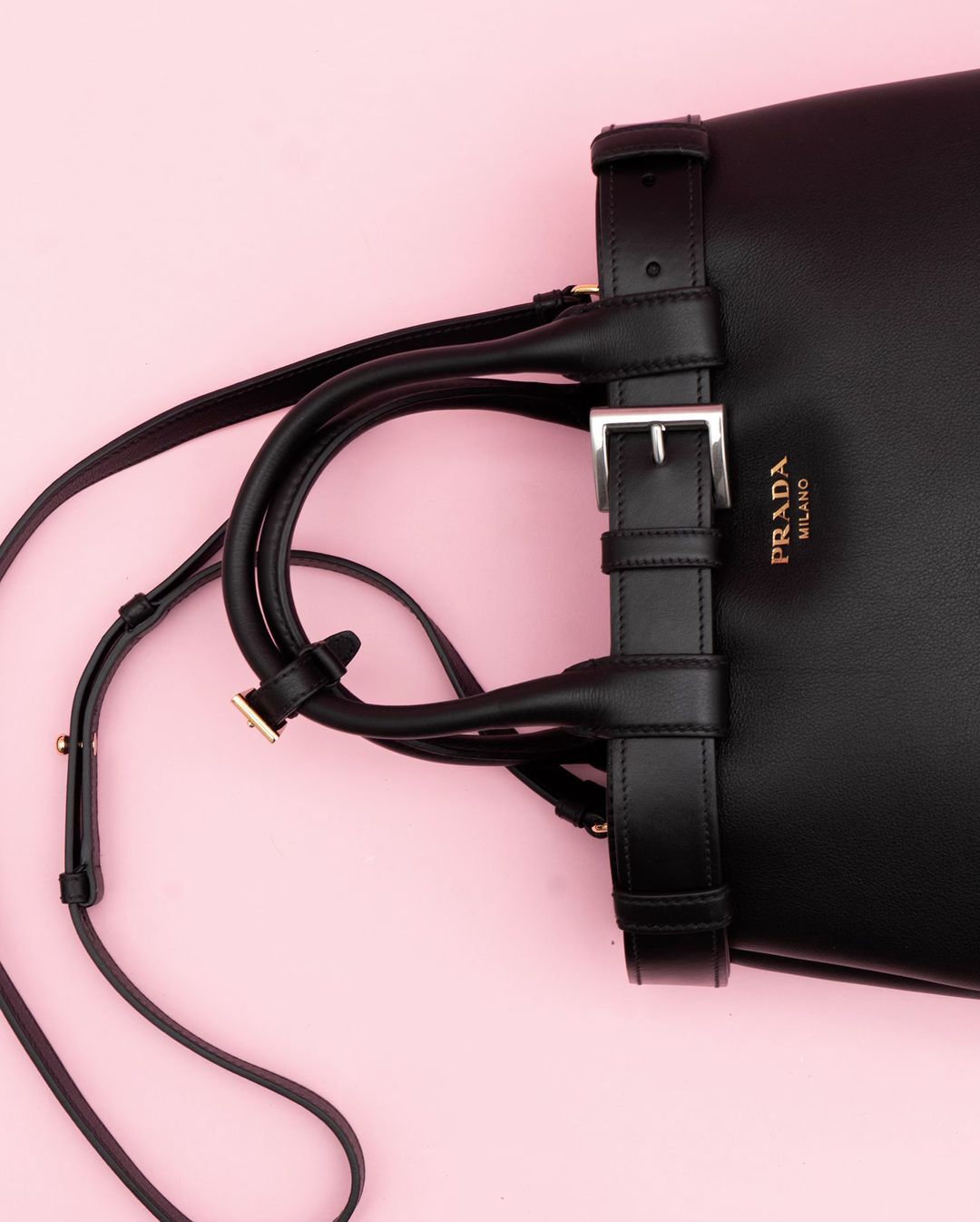 Close-up of a black leather Prada handbag with a buckle strap and shoulder strap on a light pink background, fashion accessory.
