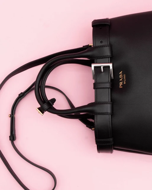 Close-up of a black leather Prada handbag with a buckle strap and shoulder strap on a light pink background, fashion accessory.