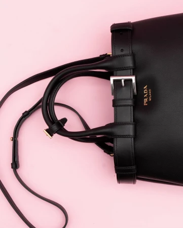 Close-up of a black leather Prada handbag with a buckle strap and shoulder strap on a light pink background, fashion accessory.