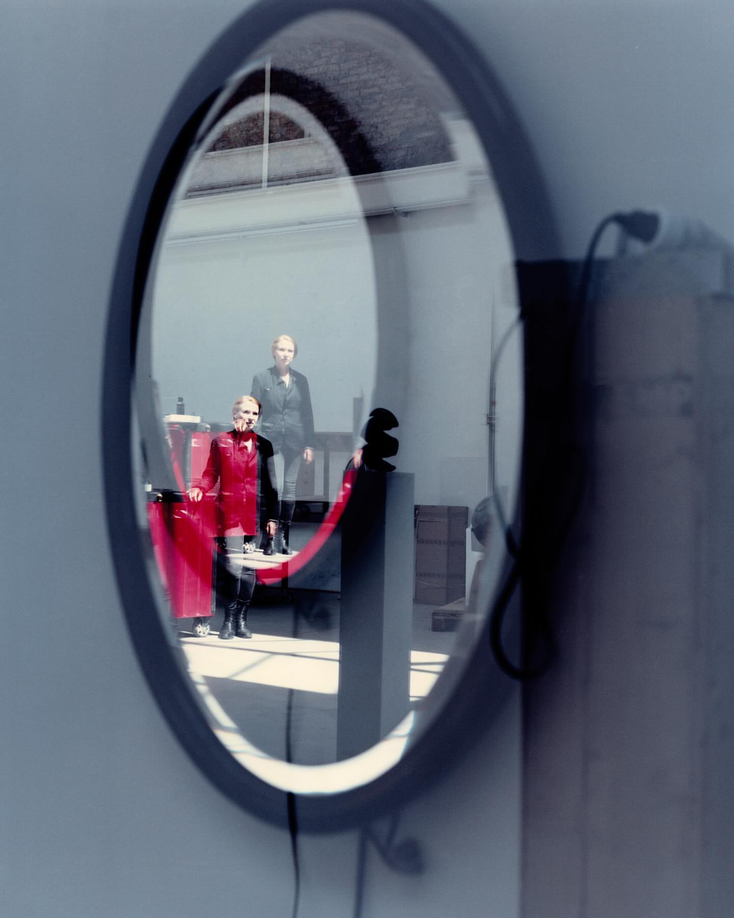 Reflection in a round mirror featuring two women in a room; one wears a red jumpsuit and the other a dark jacket.